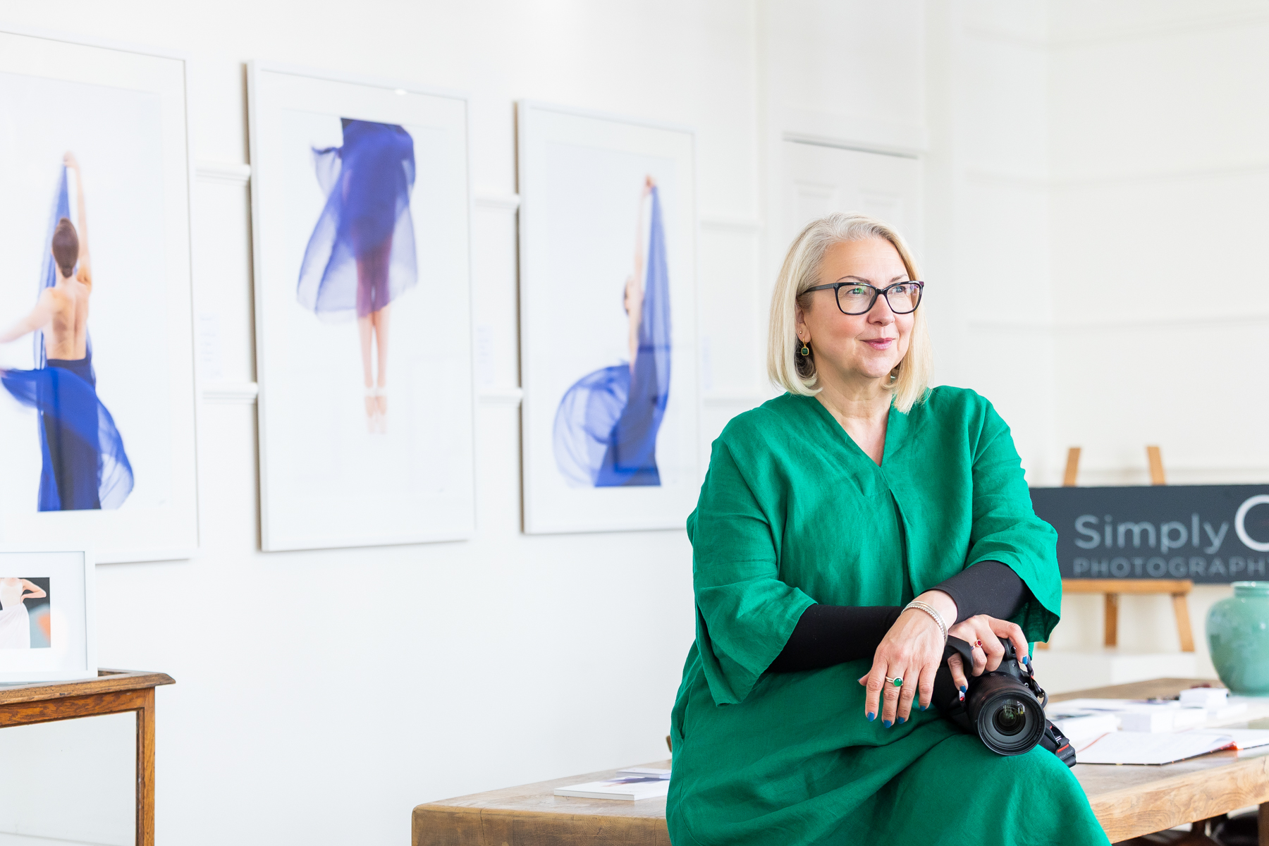Commercial branding photographer Cherry Beesley holding her camera sat on a table in natural light during her fine art exhibition in Woodbridge Suffolk looking away from the camera in a green dress sat answering questions on how to become a professional photographer